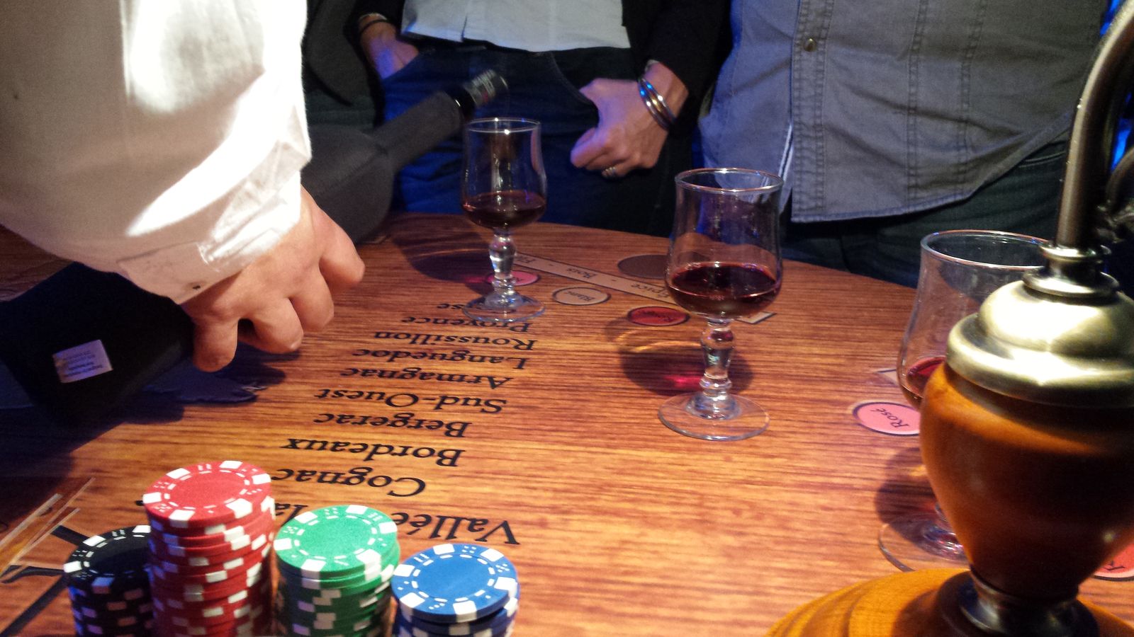 Une table de casino avec une main qui sert un verre de vin rouge. En premier plan nous avons des jetons de différentes couleurs.
