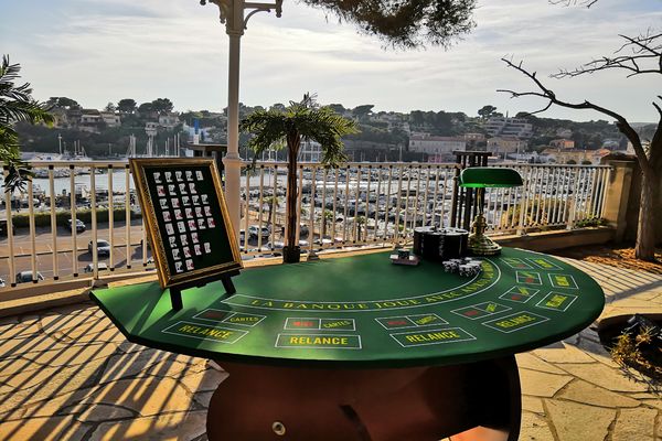 une table de casino poker devant une vue sur la mer. Une lampe, des jetons et un tableau récapitulatif des combinaisons, sont disposés sur la table.
