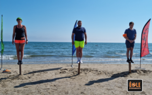trois personnes sur des poteaux à la plage avec en fond un drapeau vert puis bleu puis rouge ainsi que la mer. En bas à droite le logo d'éole événements