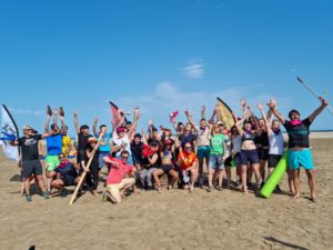 un groupe de personnes qui lèvent les bras sur la plage. Avec des objets d'aventuriers dans les mains et des drapeaux en arrière plan