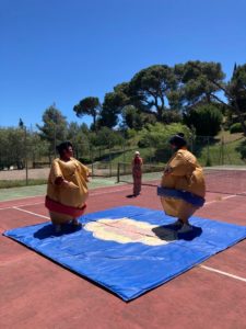 deux personnes habillés en sumos debout sur un tatami. Ils se regardent. En arrière plan une femme prend un photo les sumos.