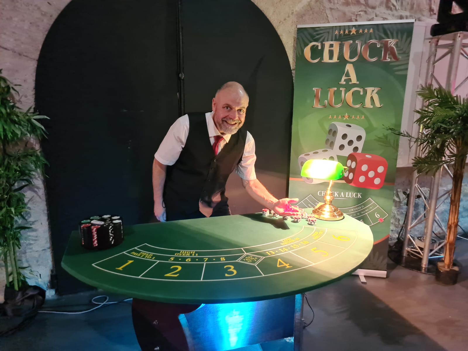 une table de casino chuck a luck avec un homme qui sourit et s'apprête à lancer des dés. En arrière plan ce trouve un roll up avec écrit chuck a luck et des dés sont affichés