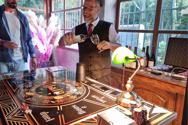 Un monsieur qui sert un verre de vin tout sourire. Il est habillé en croupier et se trouve derrière une table noir et dorée nommée Wine & song. Dessus se trouve des jetons et une lampe et derrière il y a des bouteilles de vins.