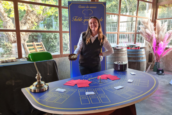 Une jeune fille habillée en croupière qui sourit et tiens un bol de fromage. Devant elle se trouve une table de casino bleu avec pour thème le casino gourmand. Dessus se trouve des serviettes, une lampe et un carrousel. Derrière elle, un roll up au même couleur que la table et une vue sur un joli extérieur avec des plantes.