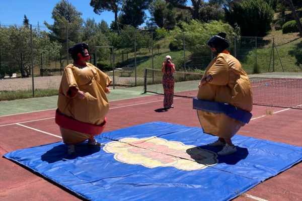 deux personnes habillés en sumos debout sur un tatami. Ils se regardent. En arrière plan une femme prend un photo les sumos.