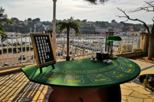une table casino poker avec dessus une petite pancarte avec des cartes, et un carrousel à côté avec une lampe. En fond il y a une terrasse et de l'eau