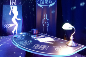 une table de casino noir avec une table, un carrousel, une tablette sur la table. En fond un roll-up noir avec un trophée dessus. Et sur la gauche un tableau james bond noir et blanc