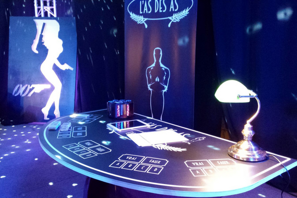 une table de casino noir avec une table, un carrousel, une tablette sur la table. En fond un roll-up noir avec un trophée dessus. Et sur la gauche un tableau james bond noir et blanc