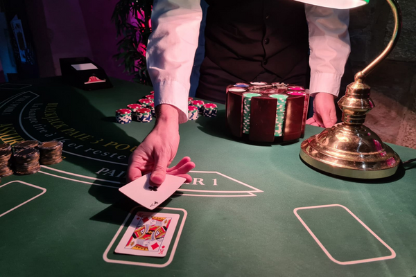 une main qui pose une carte sur une autre pour faire un blackjack. Autour de lui se trouve un carrousel, une lampe et des jetons, le tout posé sur une table de casino