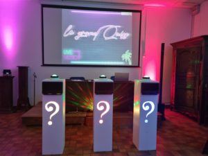 trois pupitres avec des points d'interrogation illuminés sont placés devant une table avec un ordinateur. Derrière une diapositive projetée sur le mur avec écrit "Le grand Quiz"