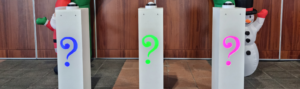 3 pupitres alignés avec des buzzers sur chacun d'eux. Les pupitres ont un point d'interrogation avec une couleur assignée, bleu, vert et rose. En fond une arche avec un père noël et un bonhomme de neige.