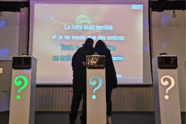 trois pupitres avec des buzzers et éclairés par des spots sont disposés devant deux personnes qui chantent devant un écran géant de karaoké.