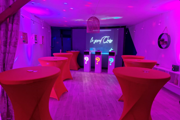 une salle aux couleurs violettes, est décorés de mange-debout aux nappes rouges avec en fond des pupitres et une diapositive projeté avec écrit "le grand quiz"