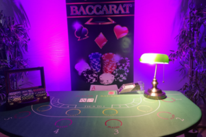 une table de casino baccarat avec dessus une boite de jetons et une lampe, en fond se trouve un roll-p up avec marqué baccarat et des lumières violettes qui décorent la pièce