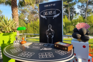 Une table de casino sur le 7ème art, elle est de couleur noir. Il y a des cases blanches avec écrit "vrai, faux, a,b,c,d" et dessus sont posés, une lampe, une caisse en bois avec des jetons ainsi qu'une décoration 007. En arrière plan, il y a des plantes, un roll up avec écrit l'as des as, des dés en guise de décoration et dessus, nous avons des ballons ainsi que des cartes posés dessus.